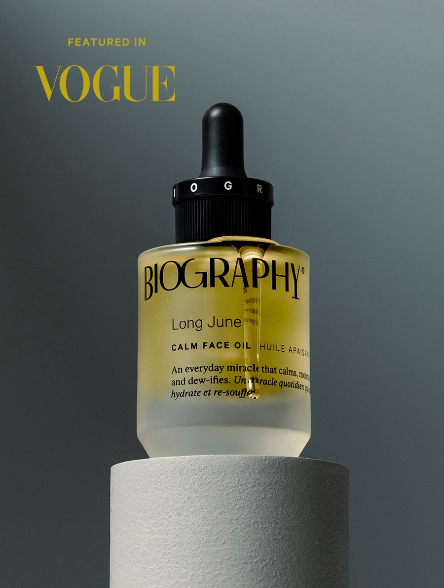Glass face oil bottle displayed on cylindrical white block, against grey background with words "FEATURED IN VOGUE" in yellow in upper left corner.