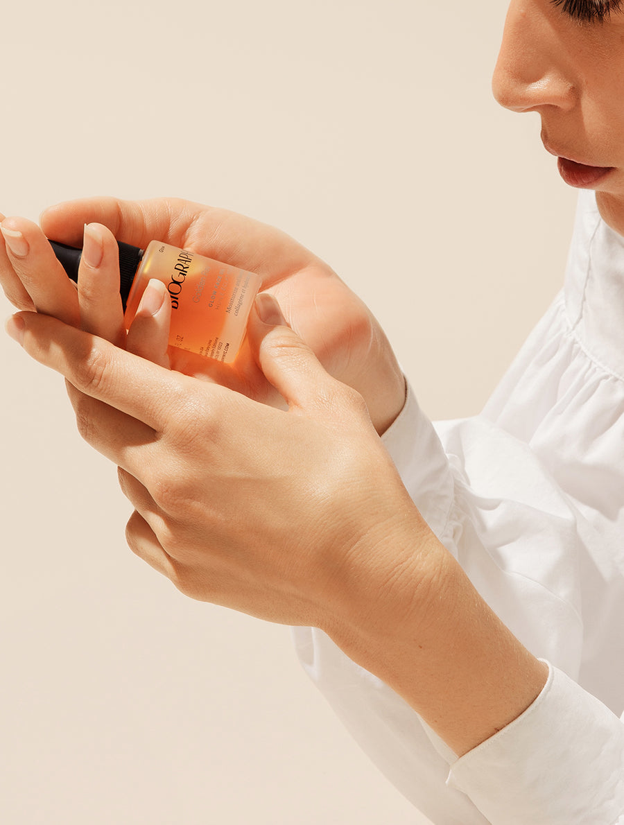 Model in white blouse holding small glass face oil bottle with both of her hands.