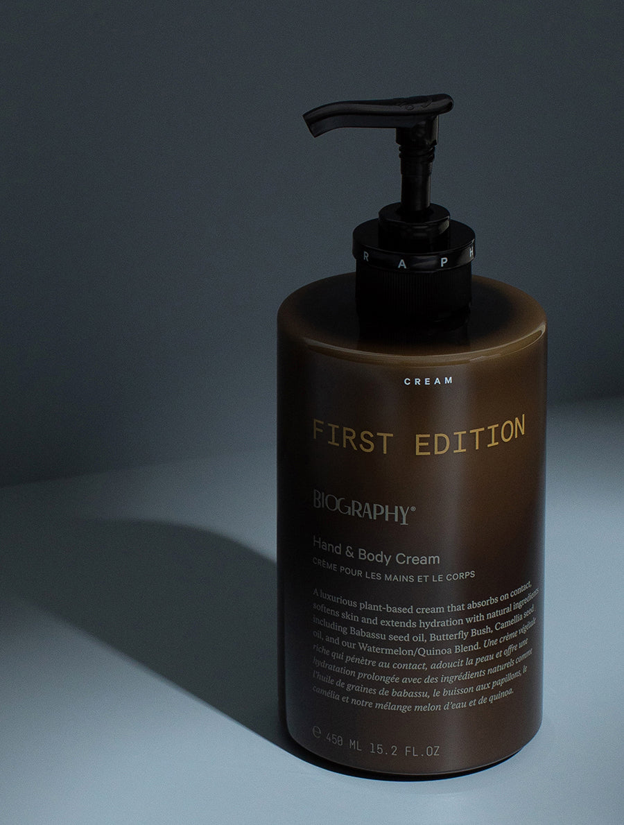 Close-up of a dark olive PCR-resin pump bottle labeled 'BIOGRAPHY First Edition Hand & Body Cream.' The bottle shows a detailed product description, mentioning its plant-based ingredients and hydration benefits. It contains 450 ml or 15.2 fl. oz of cream. Set against a muted gray background.